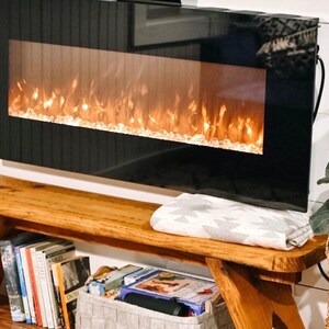 Op de afbeelding: Een houten bank met een wit deken erop, een imitatiebont kruk en een potplant met groene bladeren. De bank heeft een ingebouwde plank met boeken en een grijze opbergdoos. Een zwarte elektrische open haard met een realistisch vlammeneffect is aan de muur boven de bank gemonteerd.