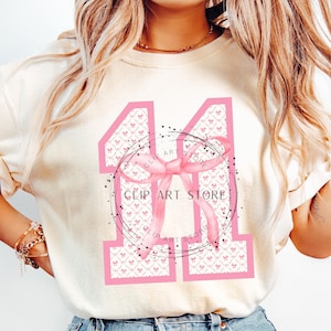Op de afbeelding: Een wit T-shirt met een roze nummer "11" gedrukt op de voorkant. Het nummer is versierd met een roze strik en een witte en roze patroonachtergrond. De tekst "Clip Art Store" is in zwart op het shirt gedrukt.