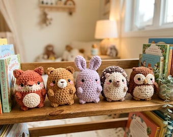 May include: A set of five crocheted plush toys arranged in a row on a wooden shelf. The figures include a rust-colored fox, a brown bear, a light purple rabbit, a cream and brown hedgehog, and a brown owl, all featuring embroidered faces.