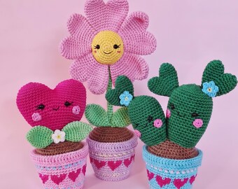 May include: Three crocheted potted plants featuring a pink flower with a yellow face, a magenta heart-shaped plant, and a green cactus with heart-shaped arms. Each plant has a smiling face, rosy cheeks, and sits in a decorative crocheted pot.