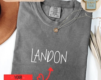 May include: A gray t-shirt with the name "LANDON" in white script. The shirt has a crew neck and short sleeves. A silver necklace with a pendant is draped across the shirt. The words "COMFORT COLORS" are printed on the shirt's tag.