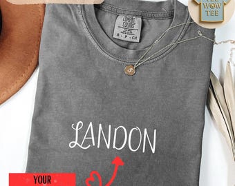 May include: A gray Comfort Colors t-shirt with the name "Landon" in white script. A silver necklace with a pendant is draped on the shirt. The shirt has a small tag that says "Comfort Colors". A red rectangle with the text "YOUR NAME HERE" is at the bottom.