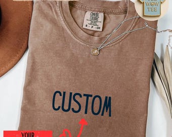 May include: A brown t-shirt with the word "CUSTOM" in navy blue. The shirt has a crew neck and a small tag that says "COMFORT COLORS". A silver necklace with a pendant is draped across the shirt. The text "YOUR NAME HERE" is in a red rectangle.
