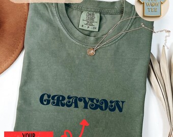 May include: An olive green t-shirt with the name "GRAYSON" in navy blue script. A silver necklace with a pendant rests on the shirt. The text "COMFORT COLORS" is at the top. A pin that says "TEE WOW TEE" is also visible.