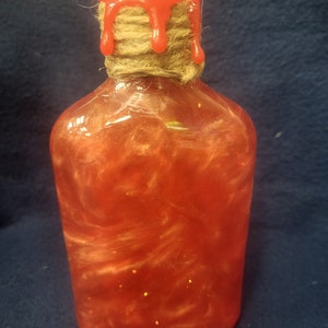 May include: A clear glass bottle with a red wax seal and a twine tied around the neck. The bottle is filled with a red liquid with gold glitter.