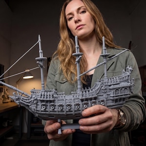 May include: A detailed gray model of a tall ship with multiple masts and intricate carvings. The model is held up by a stand and features detailed rigging and architectural elements. The ship is a miniature replica of a historical sailing vessel.
