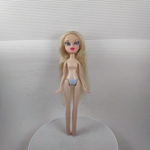 May include: A Bratz doll with long blonde hair, blue eyeshadow, and pink lipstick. The doll is wearing light blue underwear. The doll is standing on a white surface against a white background.
