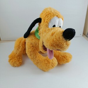 Pode incluir: Um brinquedo de pelúcia do Pluto, um cão de desenho animado da Disney. O Pluto está deitado com a língua de fora. Ele está usando uma coleira verde.