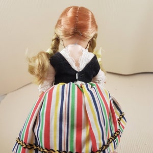 May include: A doll wearing a black vest and a colorful striped skirt with a gold trim. The doll has blonde braids and red hair.