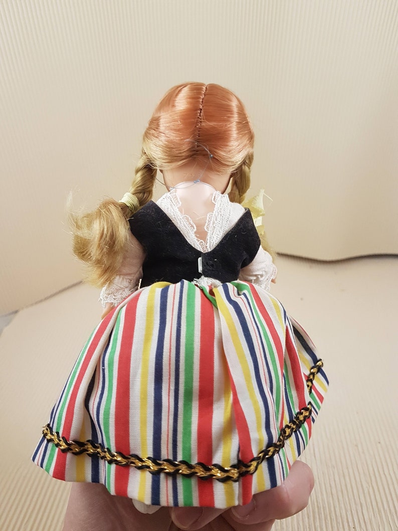 May include: A doll wearing a black vest and a colorful striped skirt with a gold trim. The doll has blonde braids and red hair.
