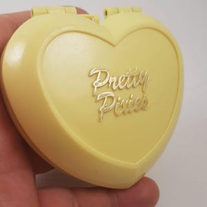 May include: A yellow heart-shaped compact with a gold inscription that reads "Pretty Pixies".