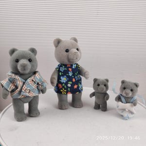 May include: Four grey bear figurines. One bear wears a blue and white checked shirt, another a blue floral dress. The other two are smaller, one standing and the other in a nappy with blue ribbons. Toy bears.