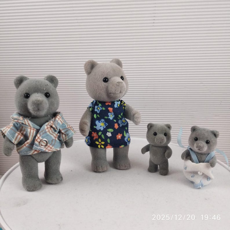 May include: Four grey bear figurines. One bear wears a blue and white checked shirt, another a blue floral dress. The other two are smaller, one standing and the other in a nappy with blue ribbons. Toy bears.