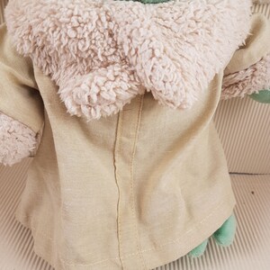 May include: A green plush toy wearing a tan coat with a white fur collar. The toy has a stitched seam down the back of the coat.