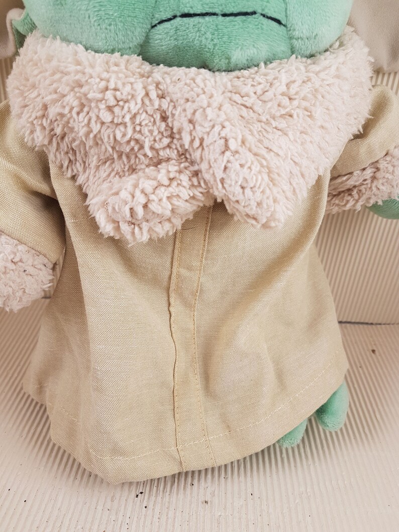 May include: A green plush toy wearing a tan coat with a white fur collar. The toy has a stitched seam down the back of the coat.