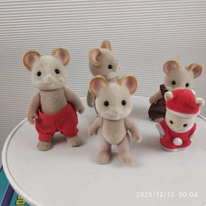 Puede incluir: Cuatro figuras de ratones en miniatura. Los ratones tienen pelaje marrón claro, orejas grandes y ojos negros. Un ratón lleva pantalones rojos, otro un atuendo marrón y un tercero está vestido con un traje rojo de Papá Noel. Las figuras están sobre una superficie blanca.