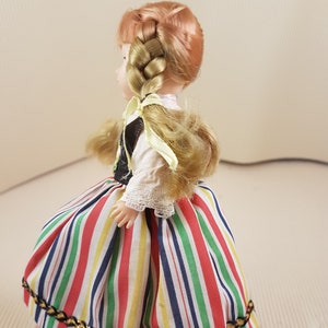 May include: A doll wearing a colorful striped skirt with a gold trim, a white blouse with lace trim, and a black vest. The doll has long blonde hair braided into a ponytail with a yellow ribbon.