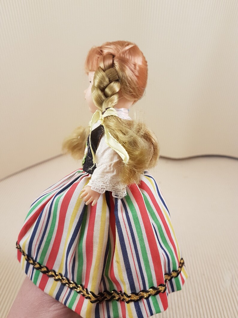 May include: A doll wearing a colorful striped skirt with a gold trim, a white blouse with lace trim, and a black vest. The doll has long blonde hair braided into a ponytail with a yellow ribbon.