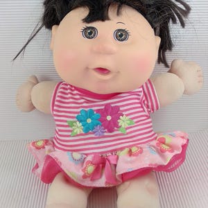 Könnte beinhalten: Eine Vintage Cabbage Patch Kids Puppe mit schwarzen Haaren und rosigen Wangen. Die Puppe trägt ein rosa-weiß gestreiftes Oberteil mit Blumenstickerei und einen rosa Blumenrock. Die Puppe sitzt.