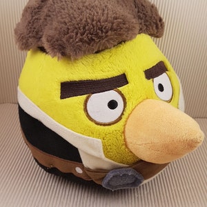 May include: Yellow plush toy of a bird with a brown fur hat. The bird has a black and white vest and a brown belt with a silver buckle. The bird has a yellow beak and white eyes.