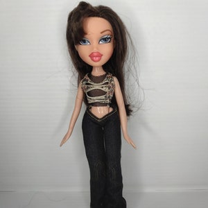 Puede incluir: Una muñeca de pelo castaño con ojos azules y labios rosas que lleva un top blanco y negro, pantalones vaqueros azules y botas marrones.
