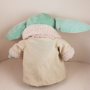 May include: A plush toy of a green character wearing a tan coat with a white fur trim. The toy has large ears and a small body.