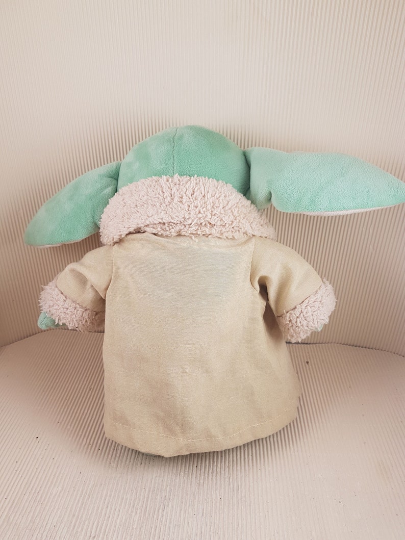 May include: A plush toy of a green character wearing a tan coat with a white fur trim. The toy has large ears and a small body.