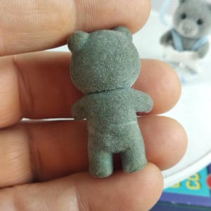 May include: A small, grey flocked teddy bear figurine, viewed from the back, with visible arms, legs, and ears. Another similar bear is visible in the background. The bear is held in a hand.