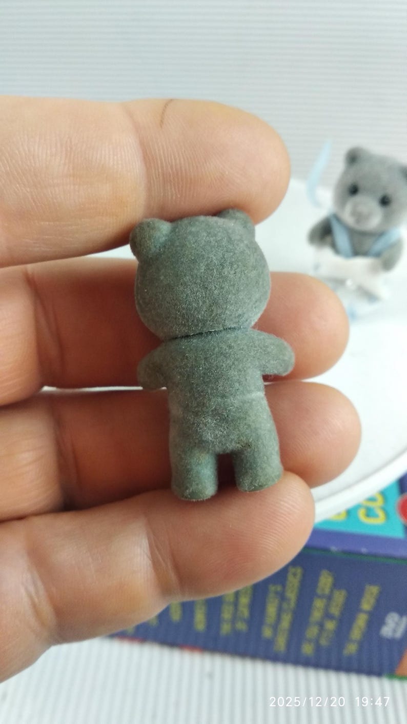 May include: A small, grey flocked teddy bear figurine, viewed from the back, with visible arms, legs, and ears. Another similar bear is visible in the background. The bear is held in a hand.