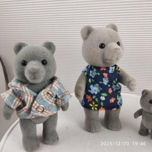 May include: Three grey bear figurines. One wears a blue and brown checked shirt. Another is in a navy dress with a floral pattern. A third, smaller bear is also visible.