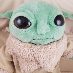 May include: A green plush toy with large eyes and a brown fur collar. The toy is wearing a tan jacket.
