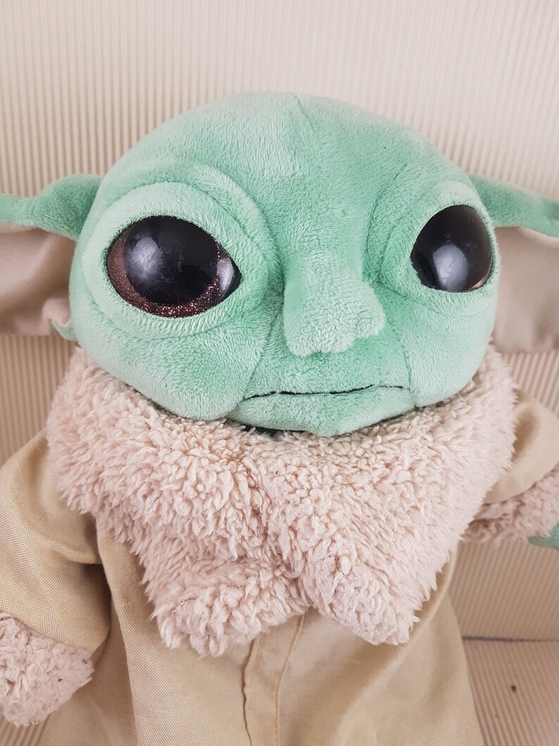 May include: A green plush toy with large eyes and a brown fur collar. The toy is wearing a tan jacket.