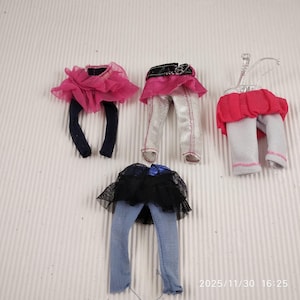 Peut inclure: Quatre ensembles de vêtements de poupée, comprenant des leggings et des jupes. Les tenues présentent une variété de couleurs, notamment rose, noir, argent et bleu. Certaines jupes sont en tulle et certaines ont des ceintures décoratives.