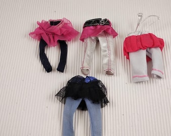 May include: Four sets of doll clothing, including leggings and skirts. The outfits feature a variety of colors, including pink, black, silver, and blue. Some skirts are made of tulle, and some have decorative belts.