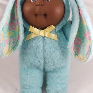 Könnte beinhalten: Vintage Cabbage Patch Kids Puppe in einem hellblauen Hasenanzug mit gelber Schleife. Der Hasenanzug hat lange Ohren mit einem floralen Muster auf der Innenseite. Die Puppe hat braune Haut und Augen.