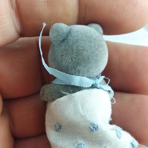 May include: A miniature grey teddy bear figure, adorned with a light blue ribbon and a white blanket featuring blue polka dots. The bear has a soft, flocked texture, a collectible item.