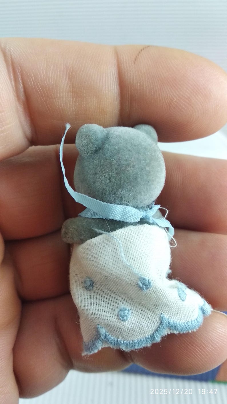 May include: A miniature grey teddy bear figure, adorned with a light blue ribbon and a white blanket featuring blue polka dots. The bear has a soft, flocked texture, a collectible item.