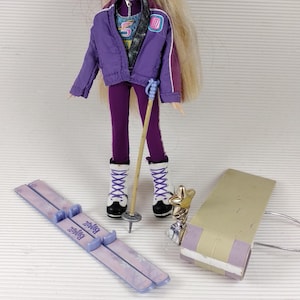 Puede incluir: Muñeca Bratz con un atuendo de esquí morado, con gafas, esquís y bastones. La muñeca tiene el pelo largo y rubio y lleva una chaqueta y pantalones morados. Los accesorios incluyen un trofeo plateado y una tabla de snowboard lila claro.