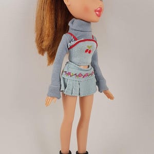 Könnte beinhalten: Eine Puppe mit langen braunen Haaren, die einen hellblauen Pullover, einen hellblauen Rock mit Blumenstickerei und schwarze Stiefel trägt. Die Puppe hat blaue Augen und rosa Lippen.