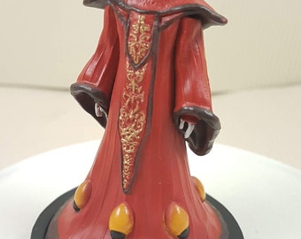 May include: A red and gold Star Wars action figure of Queen Amidala in her royal robes. The figure is standing on a black base.