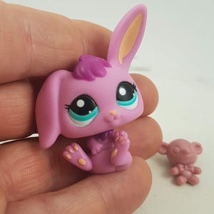May include: A pink toy bunny with large black and white eyes, teal accents, and a purple tuft of hair. The bunny is sitting with its legs extended. A small pink toy bear is also visible.