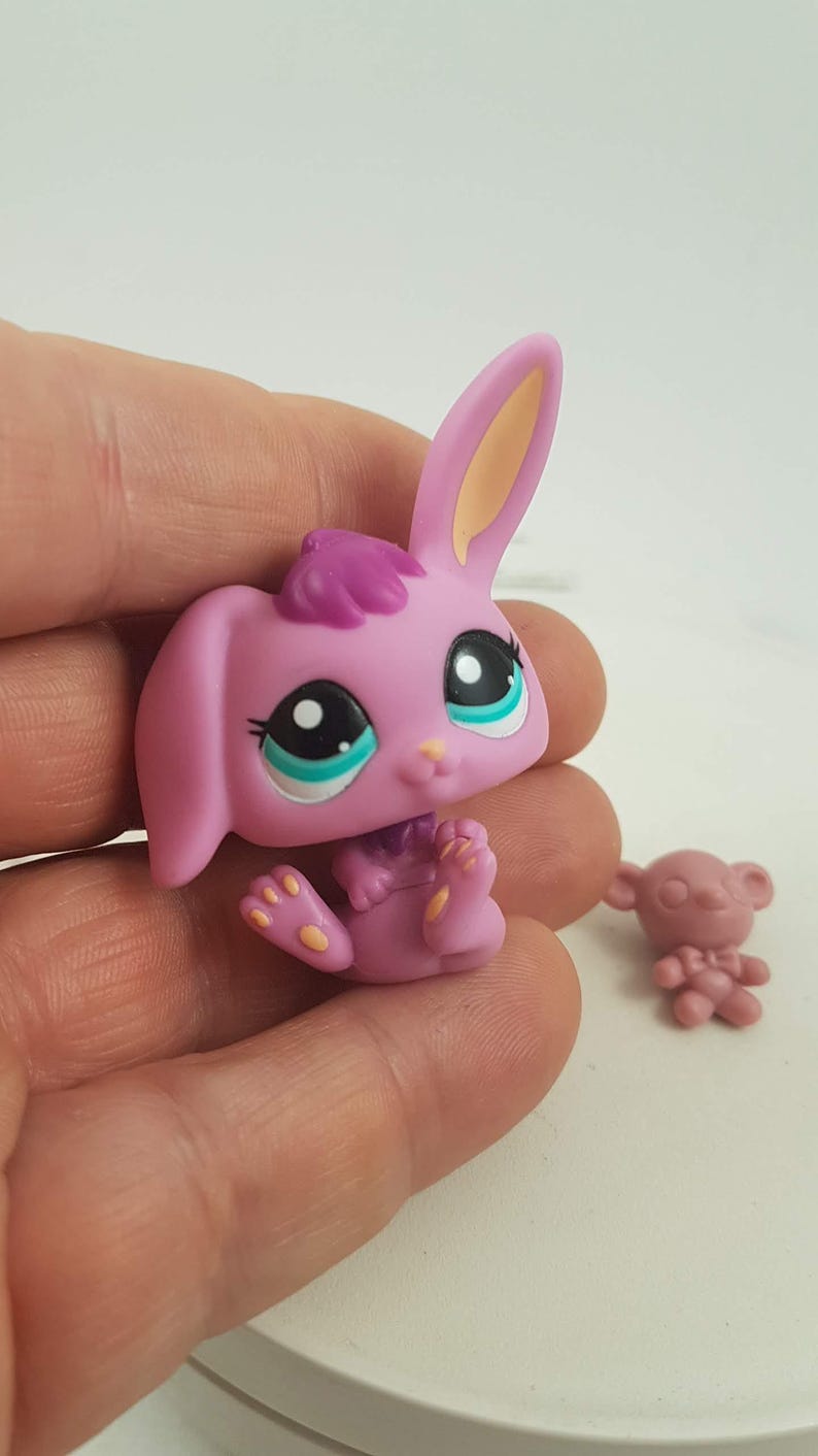 May include: A pink toy bunny with large black and white eyes, teal accents, and a purple tuft of hair. The bunny is sitting with its legs extended. A small pink toy bear is also visible.