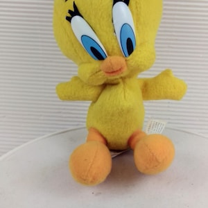 May include: A yellow plush toy of the character Tweety Bird with a large head, blue and white plastic eyes, long black eyelashes, and orange feet. The soft stuffed figure has a small tuft of yellow hair on top and outstretched arms.