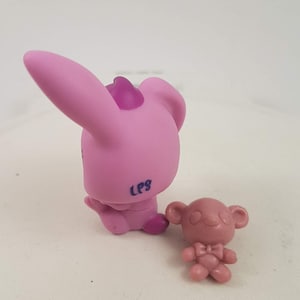 May include: Two small, pink plastic animal figurines. One is a bunny with large ears and a purple accent on its head. The other is a small bear with a bow tie. The bunny has "LPS" printed on its side.