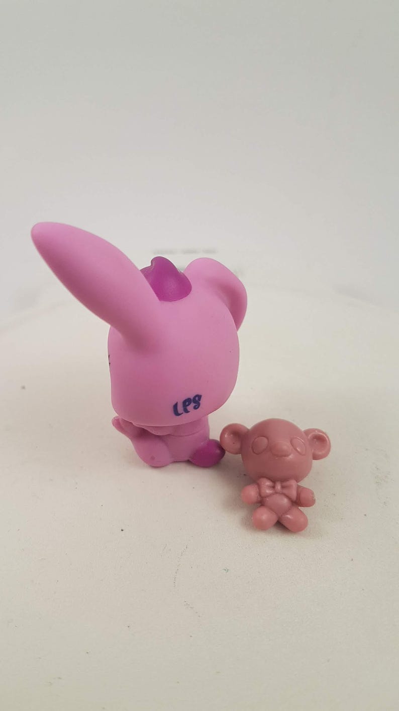 May include: Two small, pink plastic animal figurines. One is a bunny with large ears and a purple accent on its head. The other is a small bear with a bow tie. The bunny has "LPS" printed on its side.