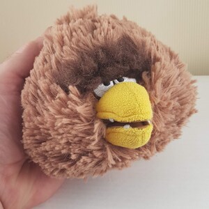 Puede incluir: Juguete de peluche marrón con un pico amarillo y dientes blancos. El juguete tiene forma de pájaro y tiene una cabeza marrón peluda.