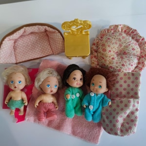 Puede incluir: Cuatro pequeños muñecos bebés con pijamas de diferentes colores y una manta de estampado rosa y blanco. Los muñecos están sentados sobre una manta de estampado rosa y blanco. Hay una pequeña silla dorada, una almohada de estampado rosa y blanco y un saco de dormir de estampado rosa y blanco en el fondo.