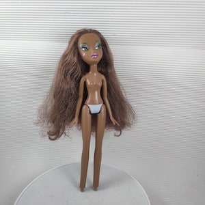 May include: A Bratz doll with long brown hair and a tan complexion. The doll has green and blue eye shadow, pink lipstick, and a star-shaped design on her cheek. She is wearing a white bikini bottom.