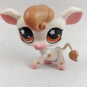 May include: A white and tan toy cow figurine with large eyes and a curly brown hair tuft. The toy has pink accents on its ears, hooves, and nose. Small star-shaped details are on its body.