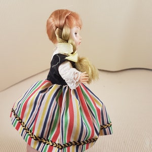 May include: A small doll with blonde hair in a braid, wearing a black bodice and a colorful striped skirt with gold trim.